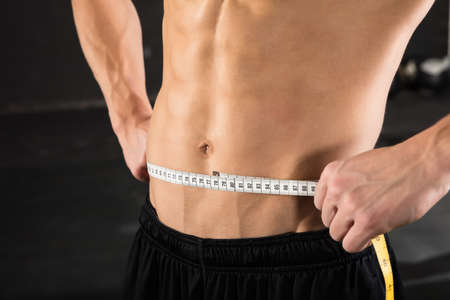 Close-up Of A Muscular Man Measuring Waist Using Measuring Tape In The Gymの写真素材