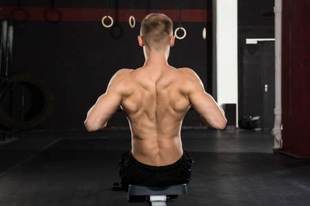 Rear View Of A Young Athlete Man Working Out On Row Machine In Gymの写真素材