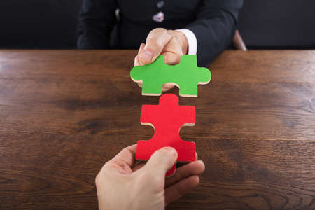 Close-up Of Two Businessmen Joining Jigsaw Pieces On Wooden Deskの写真素材
