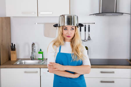 Portrait Of An Annoyed Young Woman With Cooking Pan On The Head Standing In Kitchenの写真素材