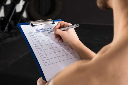 Close-up Of A Person Hand Writing A Workout Plan On Clipboard In The Gymの写真素材