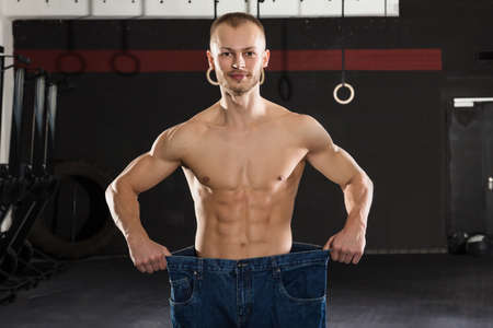 Young Athlete Man Wearing Loose Jean Showing His Weight Loss In The Gym