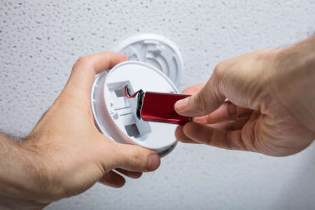 Close-up Of A Person Hand Removing Battery From Smoke Detector On Ceiling Wall