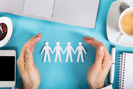 A Businessman's Hand Protecting The Paper Cut Out Human Figure On Office Deskの写真素材