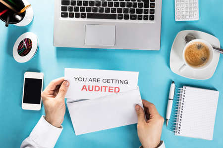 Close-up Of A Hand Holding You Are Getting Audited Card In Envelope On Office Desk