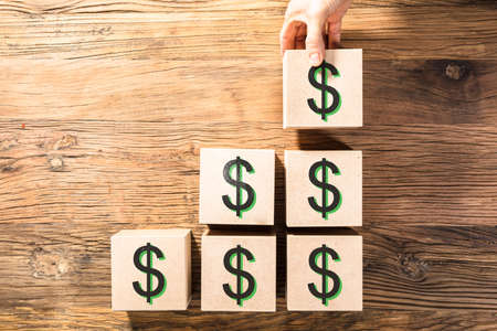 Close-up Of A Person's Hand Arranging Wooden Block With Dollar Sign On Deskの写真素材