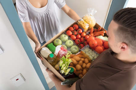 Elevated View Of Smiling Young Woman Accepting Cardboard Box Full Of Groceries From Delivery Manの写真素材