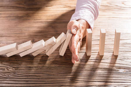 Close-up Of A Female's Hand Stopping The Domino From Falling On Wooden Deskの写真素材