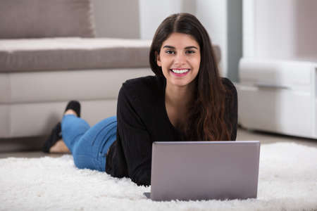 Close-up Of A Smiling Young Woman Lying On Soft Rug Using Laptop At Homeの写真素材