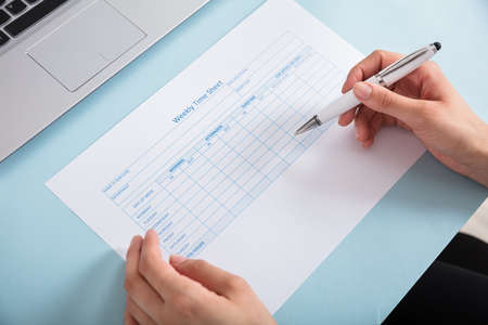 Close-up Of A Businesswoman Filling The Weekly Time Sheet On Blue Deskの写真素材