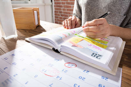 Close-up Of A Businesswoman Marking Schedule On Calendar From Diary At Workplace