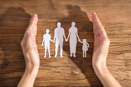 Close-up Of A Woman's Hand Protecting Family Paper Cut Out On Wooden Deskの写真素材