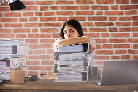 Young Businesswoman Sleeping On Folder Stacked On Office Desk At Workplaceの写真素材