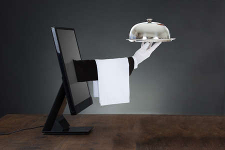 Waiter's Hand Coming Out From The Computer Screen Kept Over The Desk Against Gray Backgroundの写真素材