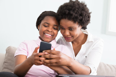 Mother And Daughter Sitting On Couch Looking At Smart Phoneのeditorial素材