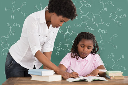 Female Teacher Assisting Student In Studies Against Green Chalkboardのeditorial素材