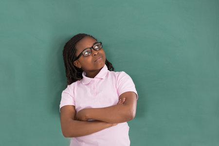 Close-up Of Thoughtful African Girl Standing Against The Green Chalkboard In Classroomのeditorial素材