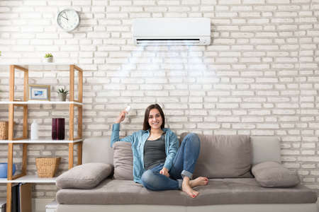 Happy Young Woman Holding Remote Control Relaxing Under The Air Conditionerの写真素材