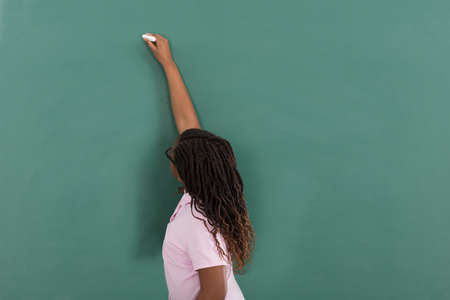 Girl Writing With Chalk On Green Chalkboard In Classroom