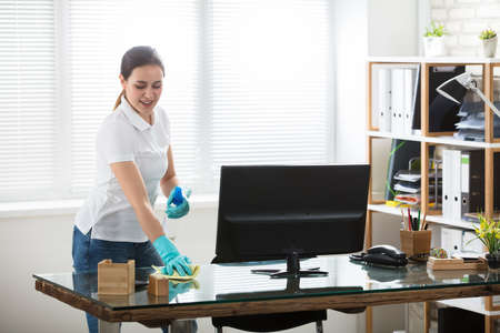 Smiling Young Woman Cleaning The Glass Desk With Rag And Spray In Officeの写真素材