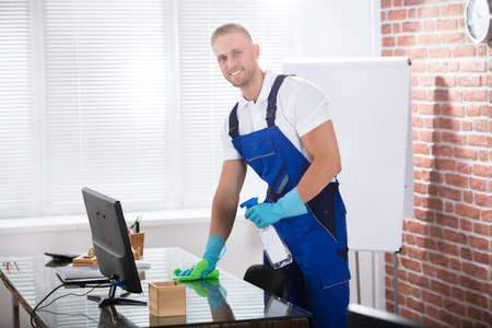 Portrait Of A Smiling Male Janitor Cleaning Desk With Cloth At Workplace