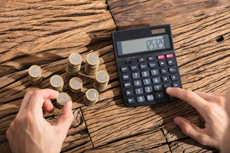 Close-up Of A Person Calculating Coins Through Calculator On Wooden Deskの写真素材