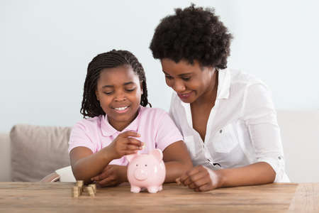 An African Girl Sitting With Her Mother Inserting Coins In Pink Piggy Bank At Homeの写真素材