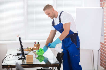 Portrait Of A Smiling Male Janitor Cleaning Desk With Cloth At Workplaceの写真素材
