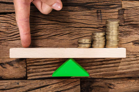 Closeup of finger balancing stacked coins on seesaw against woodの写真素材
