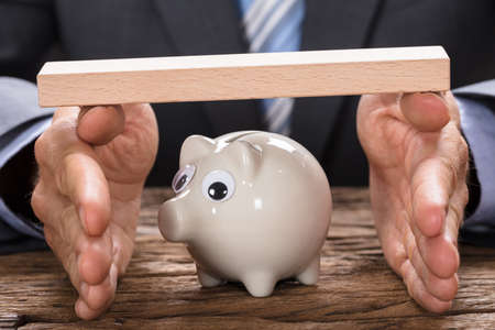 Midsection of businessman covering piggy bank with hands and wooden plank at table