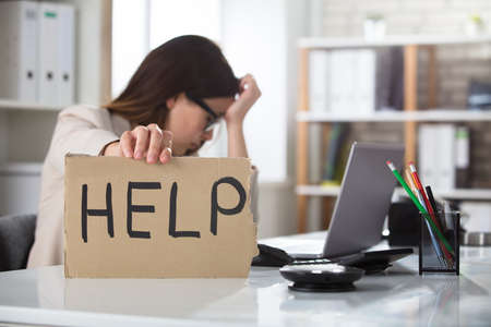 A Depressed Businesswoman Holding Help Sign On Desk At Workplace In Officeの写真素材