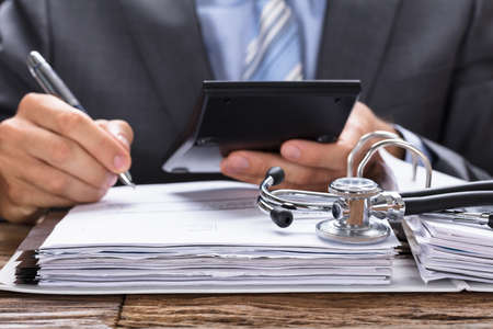 Midsection of businessman calculating invoice with stethoscope on documents at table