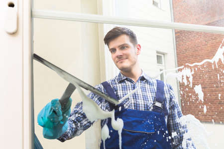 Young Male Housekeeper In Uniform Cleaning Window With Squeegee
