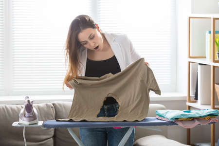 Shocked Young Woman Looking At Burnt T-shirt While Standing By Ironing Board At Homeの写真素材