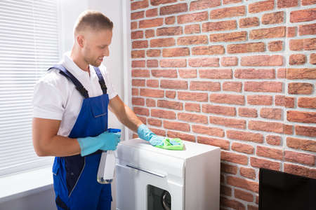 Happy Male Janitor In Uniform Cleaning The Furniture With Rag And Spray