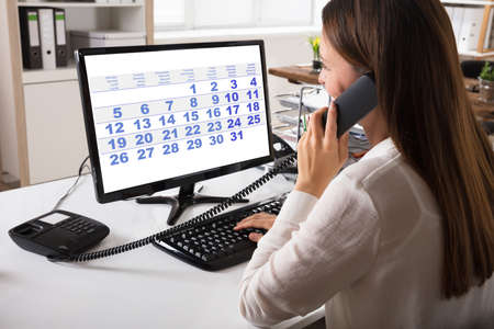Close-up Of A Young Businesswoman Talking On Landline Phone With Calendar On Computer Screenの写真素材