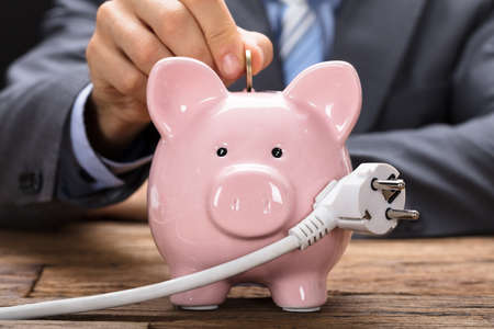 Midsection of businessman putting coin in piggybank with electric plug at wooden tableの写真素材