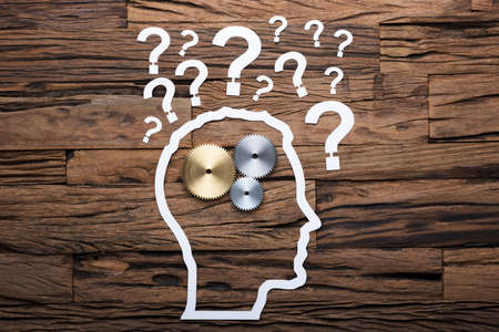 Paper businessman's head outline with metallic gears and question marks on wooden table