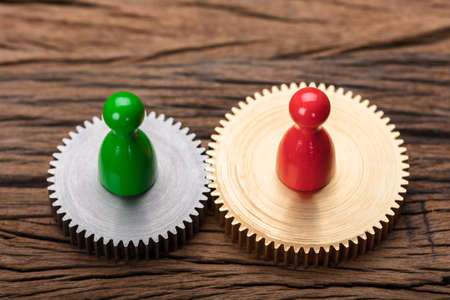 Closeup of red and green pawn figurines on interlocked cogwheels on wooden tableの写真素材