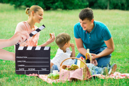 Hands holding clapperboard while young family enjoying picnic on grassy fieldのeditorial素材