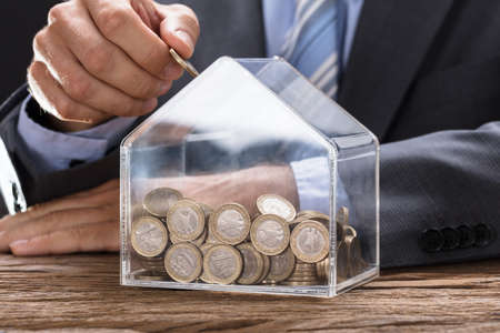 Midsection of businessman putting coin in transparent house shaped piggybank at wooden table