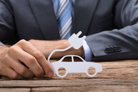 Midsection of businessman holding paper electric car at wooden tableの写真素材
