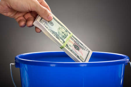 Cropped hand of businessman throwing US paper currency bundle in blue bucket against gray backgroundの写真素材