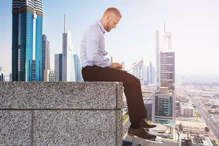 Young Businessman Sitting On The Edge Of High Roof Using Laptopの写真素材