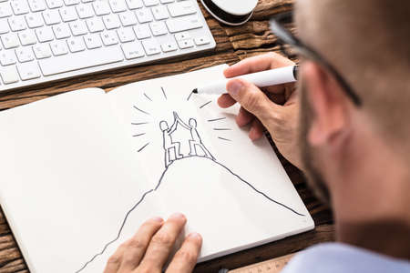 Businessman Drawing The Figures Giving High Five On Top Of Mountain In Notebookの写真素材