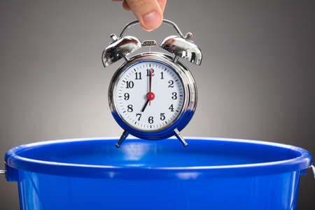 Cropped hand of businessman throwing alarm clock in blue bucket against gray backgroundの写真素材