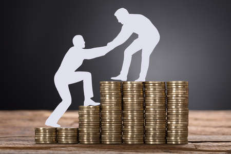 Closeup of paper businessman pulling colleague while standing on stacked coins against black...