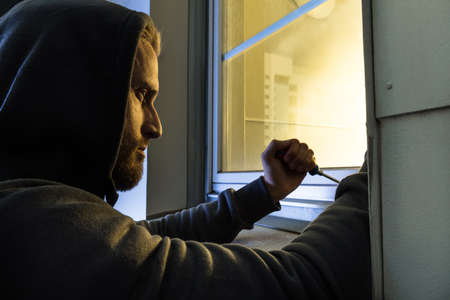 Male Young Robber In Hood Using The Screwdriver To Open The Glass Windowの写真素材