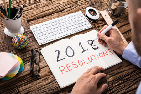 Elevated View Of Businessman Writing 2018 Resolutions On Notebook At Workplace