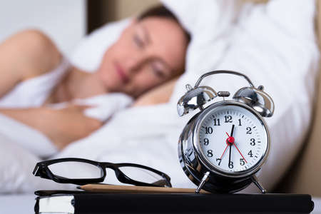 Close-up Of Eyeglasses And Alarm Clock On Table In Front Of Sleeping Woman